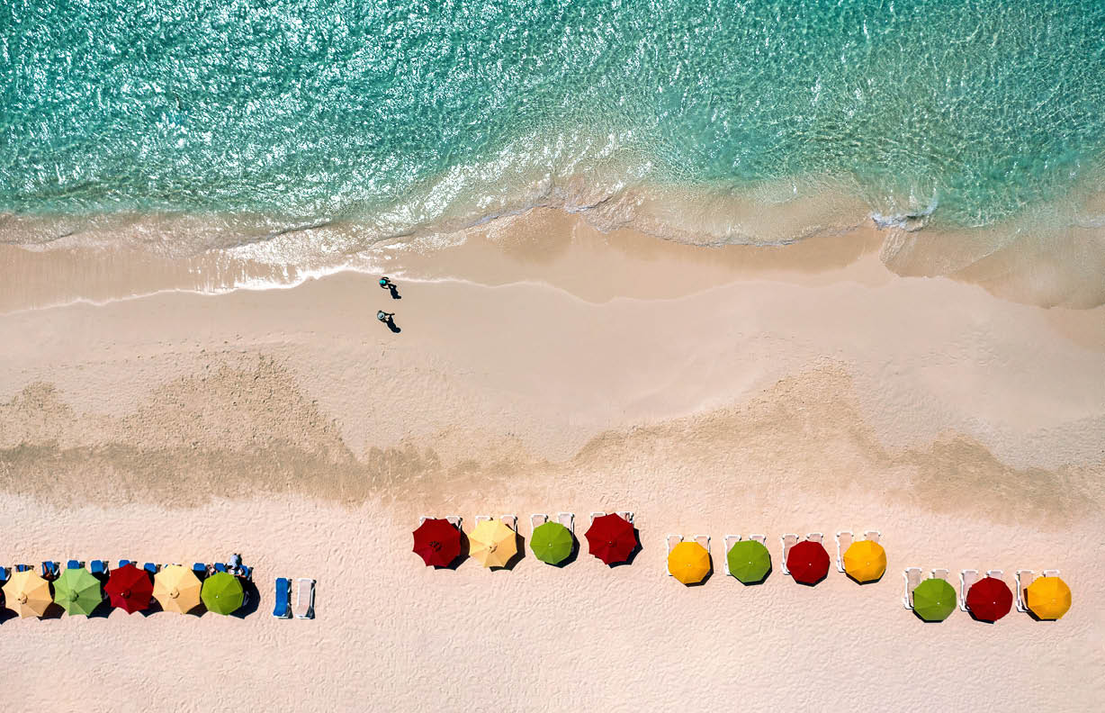 Aerial view of colorful umbrellas along a beach with two walkers on Rendezvous Bay in Anguilla.