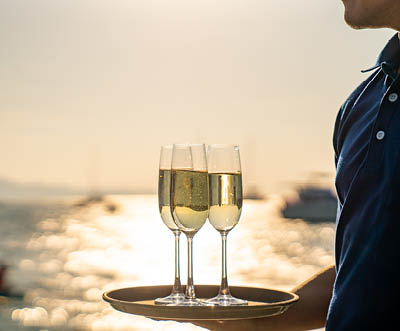 Asian man waiter holding champagne glass on the tray serving to group of passenger tourist travel on luxury catamaran boat yacht sailing in the ocean at summer sunset on beach holiday vacation trip.