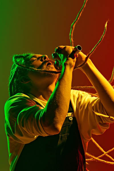 Dynamic image of artistic, soulful young man with dreadlocks, musician singing solo against gradient red green background in neon light. Concept of music, performance, festival, concert