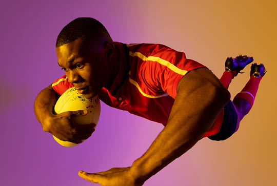 African american male rugby player jumping with rugby ball over pink lighting. Sport, movement, training and active lifestyle concept.