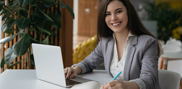 Portrait of beautiful confident businesswoman using laptop computer taking notes working online in office. Smiling student studying looking at camera sitting modern library, education concept