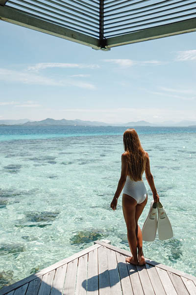 Back view young woman traveler stand on wooden pier holding flippers ready for snorkeling in blue ocean. Summer by the sea concept. Young woman in bikini going to snorkeling in a sea