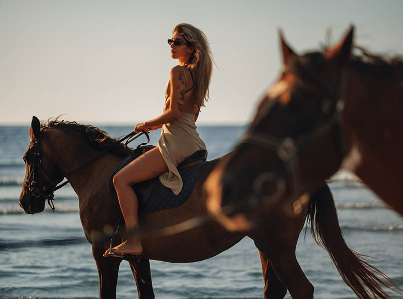 Tattooed beautiful young woman horseback riding on coastline at the beach during a sunset. Portrait of a female in a backless dress enjoying freedom at the seacoast with her horse.
