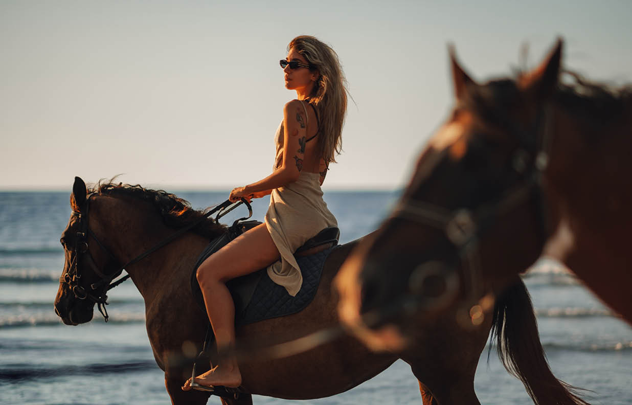 Tattooed beautiful young woman horseback riding on coastline at the beach during a sunset. Portrait of a female in a backless dress enjoying freedom at the seacoast with her horse.