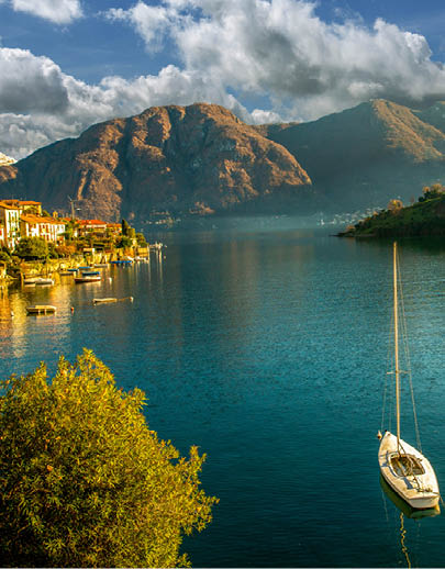 Idyllic landscape with white yacht (nautical vessel) next to coast on lake Como /At distant house , on other side - mountains and snow-capped also. Houses also on other side and Church on island.