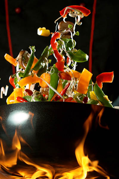 Closeup of chef throwing vegetable mix from wok pan in fire. Fresh asian food preparation on dark background.
