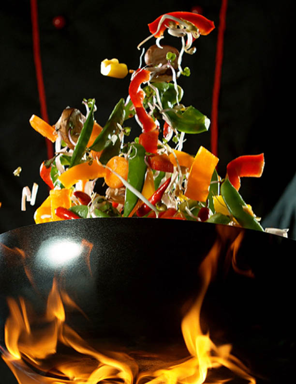 Closeup of chef throwing vegetable mix from wok pan in fire. Fresh asian food preparation on dark background.