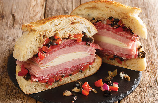 New Orleans Muffuletta Sandwich a monster of a sandwich piled high with multiple types of meat and cheese, and a perky olive salad close up on a slate board on the table. Horizontal