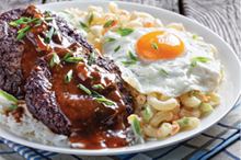loco moco of white rice, topped with a hamburger patty, a fried egg, brown gravy, and macaroni salad served on a white plate on a rustic wooden table, Hawaiian cuisine, view from above, 