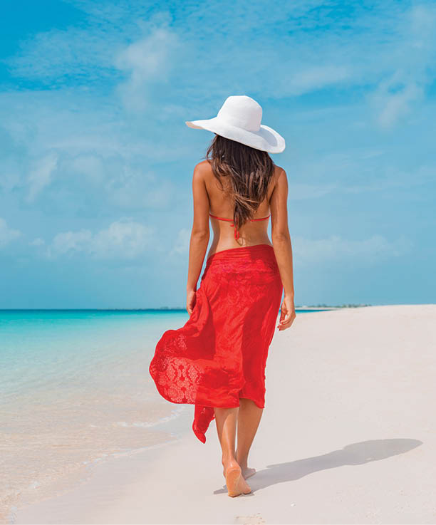 Luxury beach vacation elegant tourist woman walking relaxing in red beachwear and sunhat on white sand Caribbean beach. Lady tourist on holiday vacation resort.
