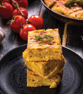 Slice of tortilla de patatas on a black plate. Traditional spanish dish with garlic sauce aioli and fresh tomatoes cherry. Omelette with eggs, potatoes and onion. Rustic black background