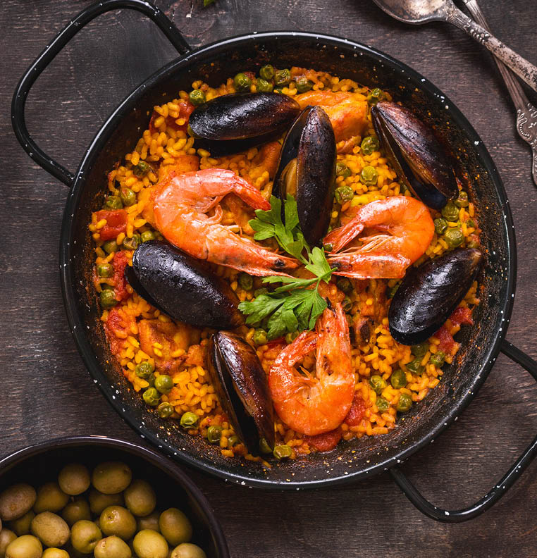 Paella in black pan with rice, shrimps, mussels, squid and meat, bowl with olives and vintage cutlery. Seafood paella, traditional spanish dish. Paella on rustic table. Selective focus. Top view