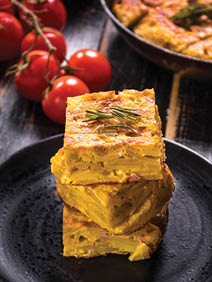 Slice of tortilla de patatas on a black plate. Traditional spanish dish with garlic sauce aioli and fresh tomatoes cherry. Omelette with eggs, potatoes and onion. Rustic black background