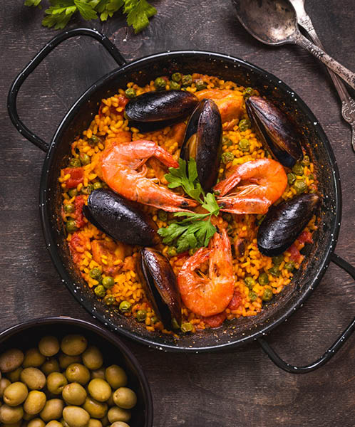 Paella in black pan with rice, shrimps, mussels, squid and meat, bowl with olives and vintage cutlery. Seafood paella, traditional spanish dish. Paella on rustic table. Selective focus. Top view