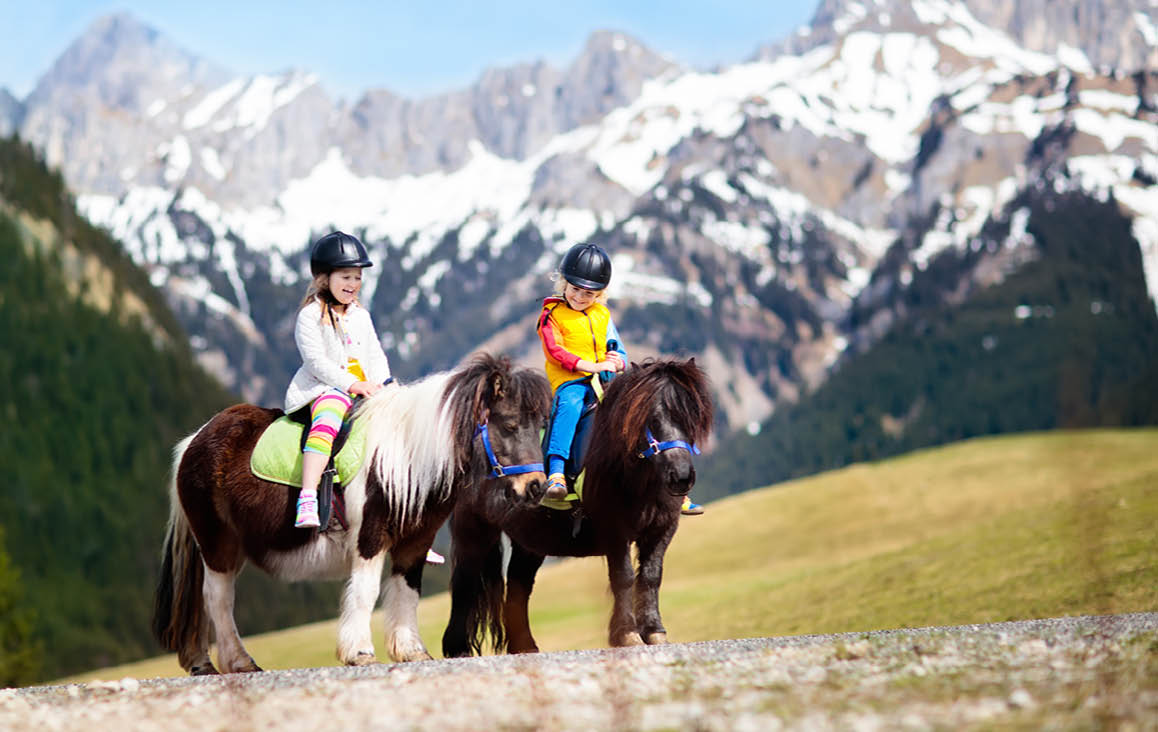 Kids riding pony in the Alps mountains. Family spring vacation on horse ranch in Austria, Tirol. Children ride horses. Kid taking care of animal. Child and pet. Little girl and boy in saddle on pony.