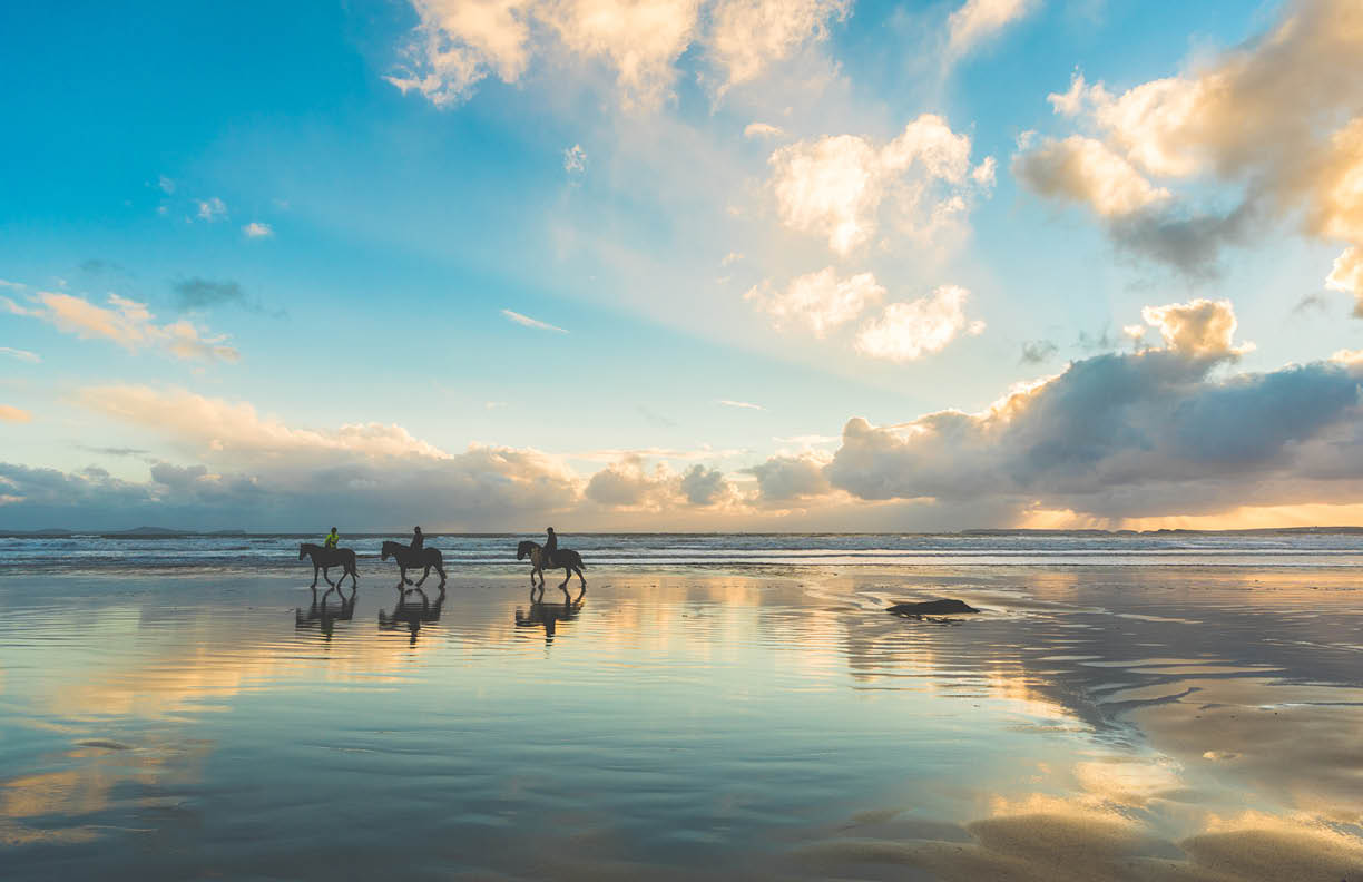 Horses walking on the beach at sunset. Three people riding horses at seaside on a cloudy day. Epic photo, wide angle shot, backlight with silhouette. Sport and travel concepts