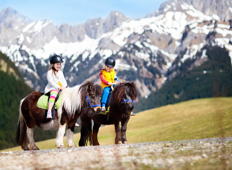 Kids riding pony in the Alps mountains. Family spring vacation on horse ranch in Austria, Tirol. Children ride horses. Kid taking care of animal. Child and pet. Little girl and boy in saddle on pony.