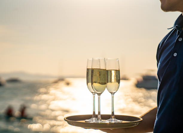 Asian man waiter holding champagne glass on the tray serving to group of passenger tourist travel on luxury catamaran boat yacht sailing in the ocean at summer sunset on beach holiday vacation trip.