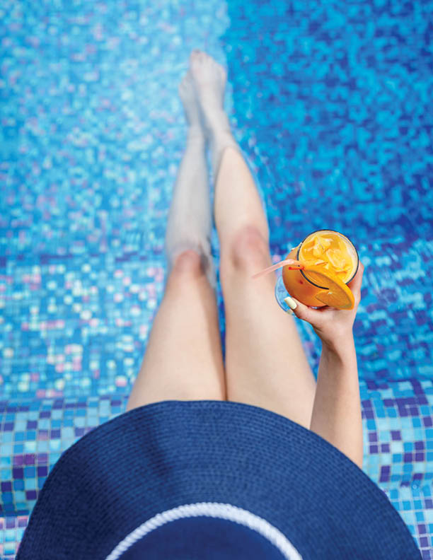 Woman in a blue summer hat with slim legs relaxing at the pool with a  cocktail 