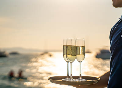 Asian man waiter holding champagne glass on the tray serving to group of passenger tourist travel on luxury catamaran boat yacht sailing in the ocean at summer sunset on beach holiday vacation trip.