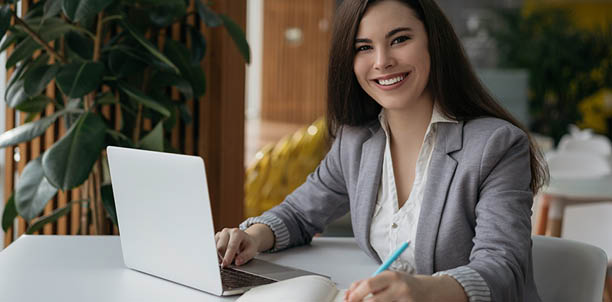 Portrait of beautiful confident businesswoman using laptop computer taking notes working online in office. Smiling student studying looking at camera sitting modern library, education concept
