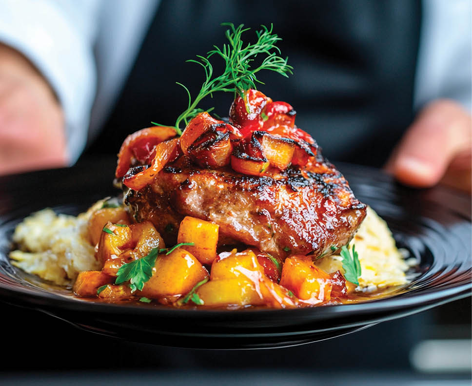 A skilled chef showcases an exquisite dish featuring succulent grilled meat, adorned with a colorful sauce and fresh herbs. The presentation highlights culinary artistry in a modern kitchen setting.
