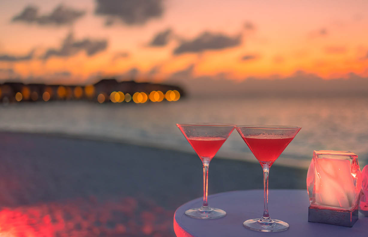 Two cocktail drinks blur beach coast party people and colorful sunset sky in background. Luxury outdoor leisure lifestyle, relaxing and romantic colors, blurred people partying on a summer evening