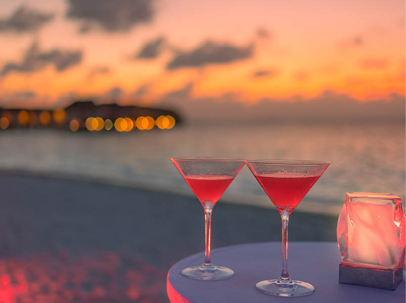 Two cocktail drinks blur beach coast party people and colorful sunset sky in background. Luxury outdoor leisure lifestyle, relaxing and romantic colors, blurred people partying on a summer evening