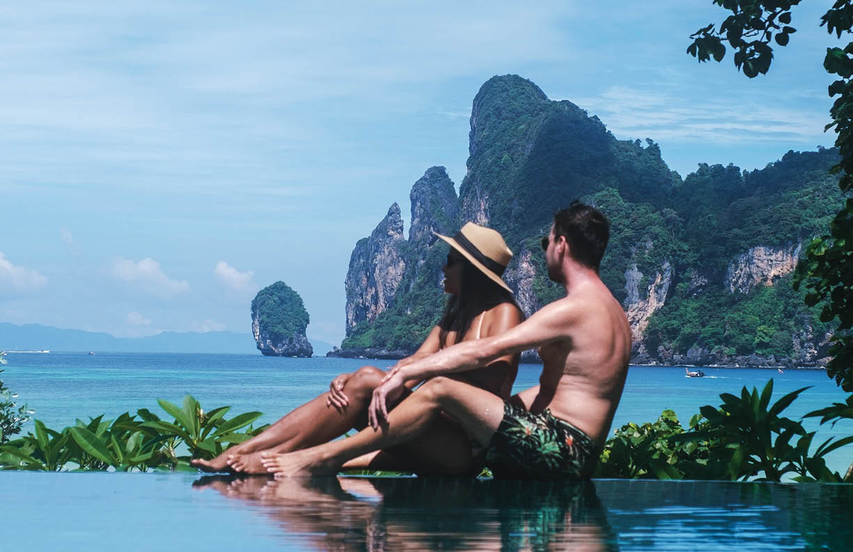 A couple relaxes at an infinity pool, savoring their holiday in Koh Phi Phi, Thailand. They enjoy breathtaking views of towering limestone cliffs and vibrant turquoise waters, embodying luxury travel.