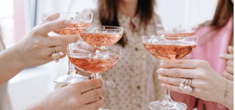 Close-up of elegant female hands clinking coupe glasses filled with pink champagne and floral garnishes during a joyful festive celebration.