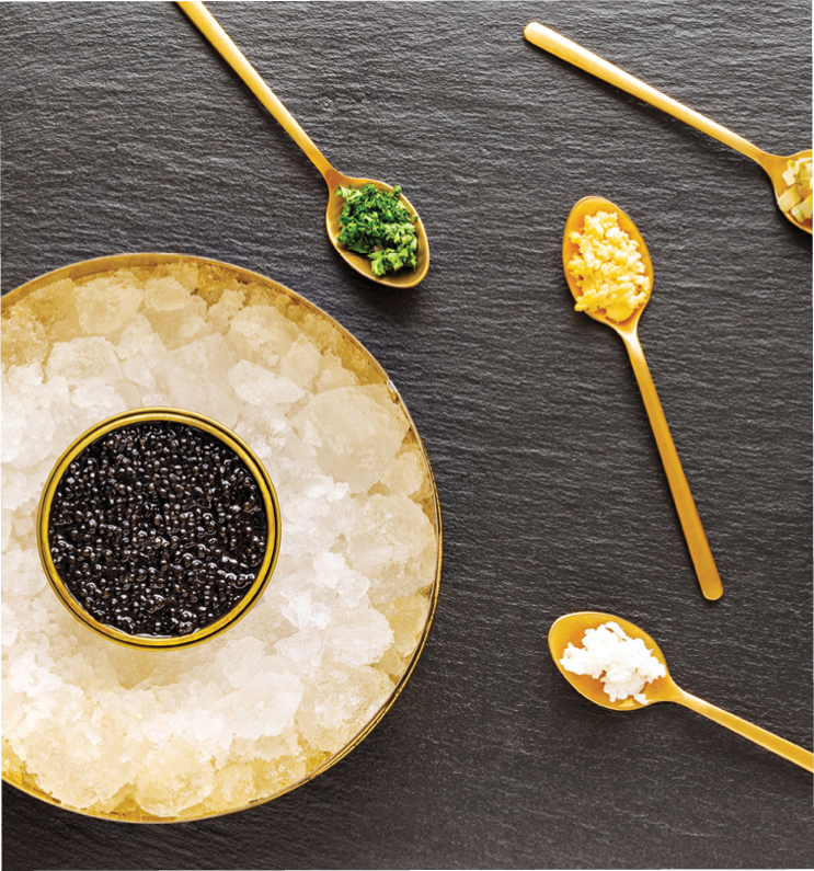 High quality real black sturgeon caviar in a metal golden box on crushed ice aside golden spoons with pickles, red onions, egg, and herbs on a slate background. Flat Lay. Top view.