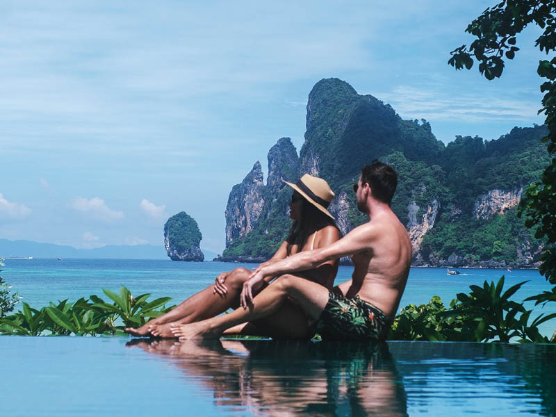 A couple relaxes at an infinity pool, savoring their holiday in Koh Phi Phi, Thailand. They enjoy breathtaking views of towering limestone cliffs and vibrant turquoise waters, embodying luxury travel.