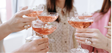 Close-up of elegant female hands clinking coupe glasses filled with pink champagne and floral garnishes during a joyful festive celebration.