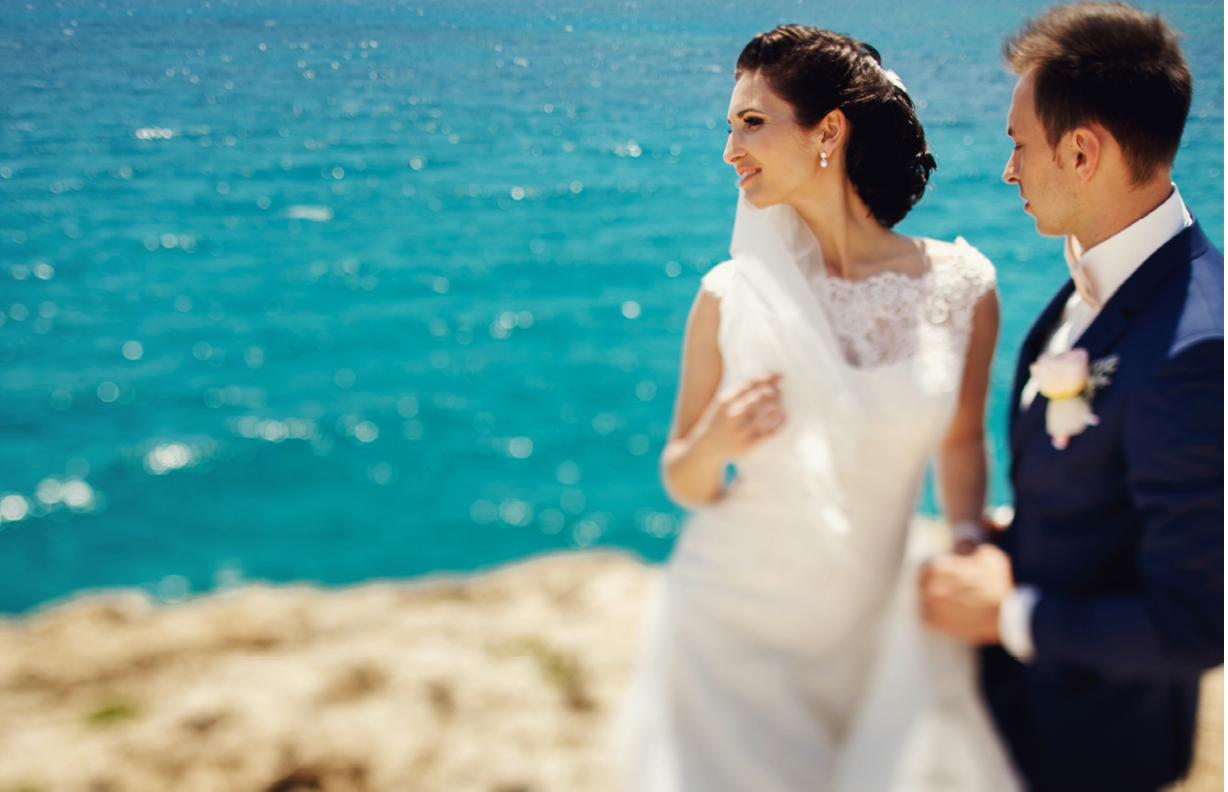 Elegant smiling young bride and groom walking on the beach, kissing and having fun, wedding ceremony near the rocks and ocean