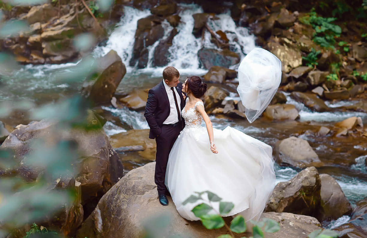 young wedding couple of sexy pretty girl or woman with hair in white bride dress and handsome man near beautiful grand waterfall in mountain