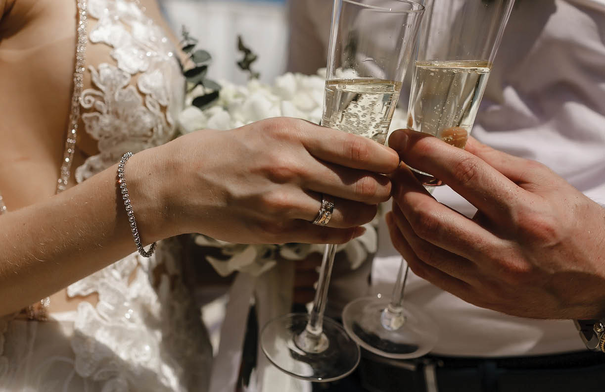 Bride and groom holding beautifully decorated wedding glasses with champaign. Wine glasses in hands on the background of wedding decorations, sunny weather.