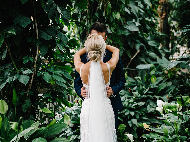 Portrait the groom and the bride standing back and kissing on the background of greenery. Wedding ceremony in Botanical green garden.