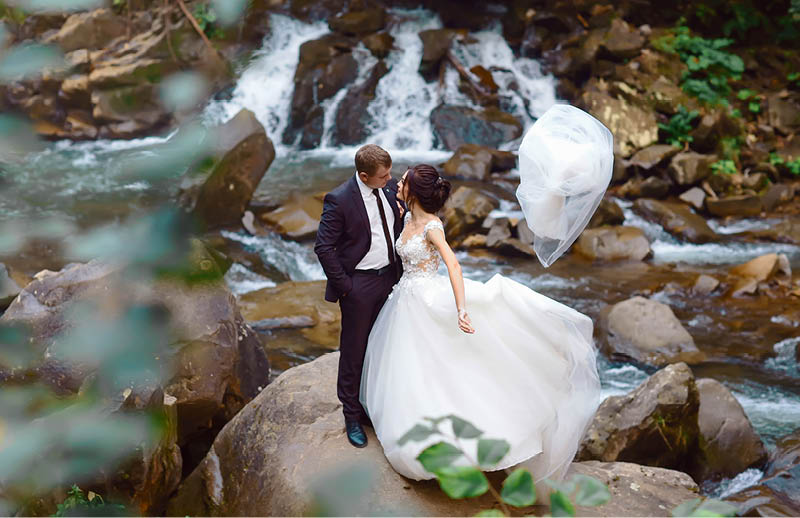 young wedding couple of sexy pretty girl or woman with hair in white bride dress and handsome man near beautiful grand waterfall in mountain