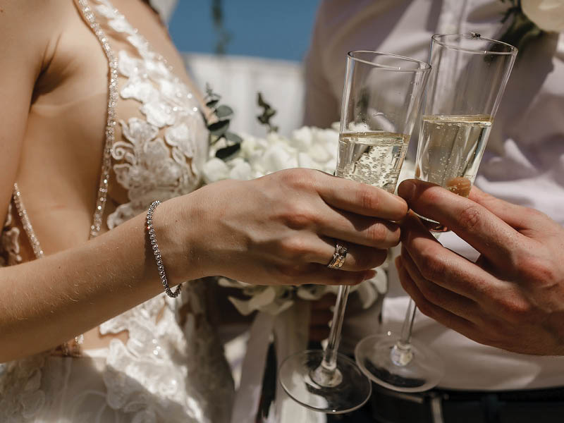 Bride and groom holding beautifully decorated wedding glasses with champaign. Wine glasses in hands on the background of wedding decorations, sunny weather.