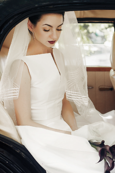 gorgeous elegant bride posing in stylish retro black car, sitting inside in saloon holding bouquet of callas. luxury wedding in vintage style. unusual gown and veil